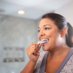 Woman brushing her teeth with clear aligners in a bright bathroom.